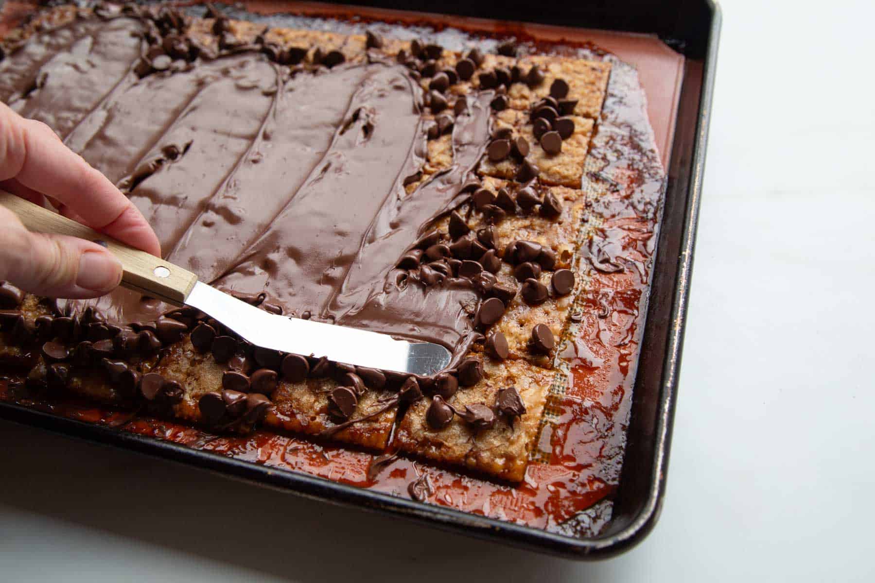 hand spreading melted chocolate chips on top of the warm caramelized saltines.