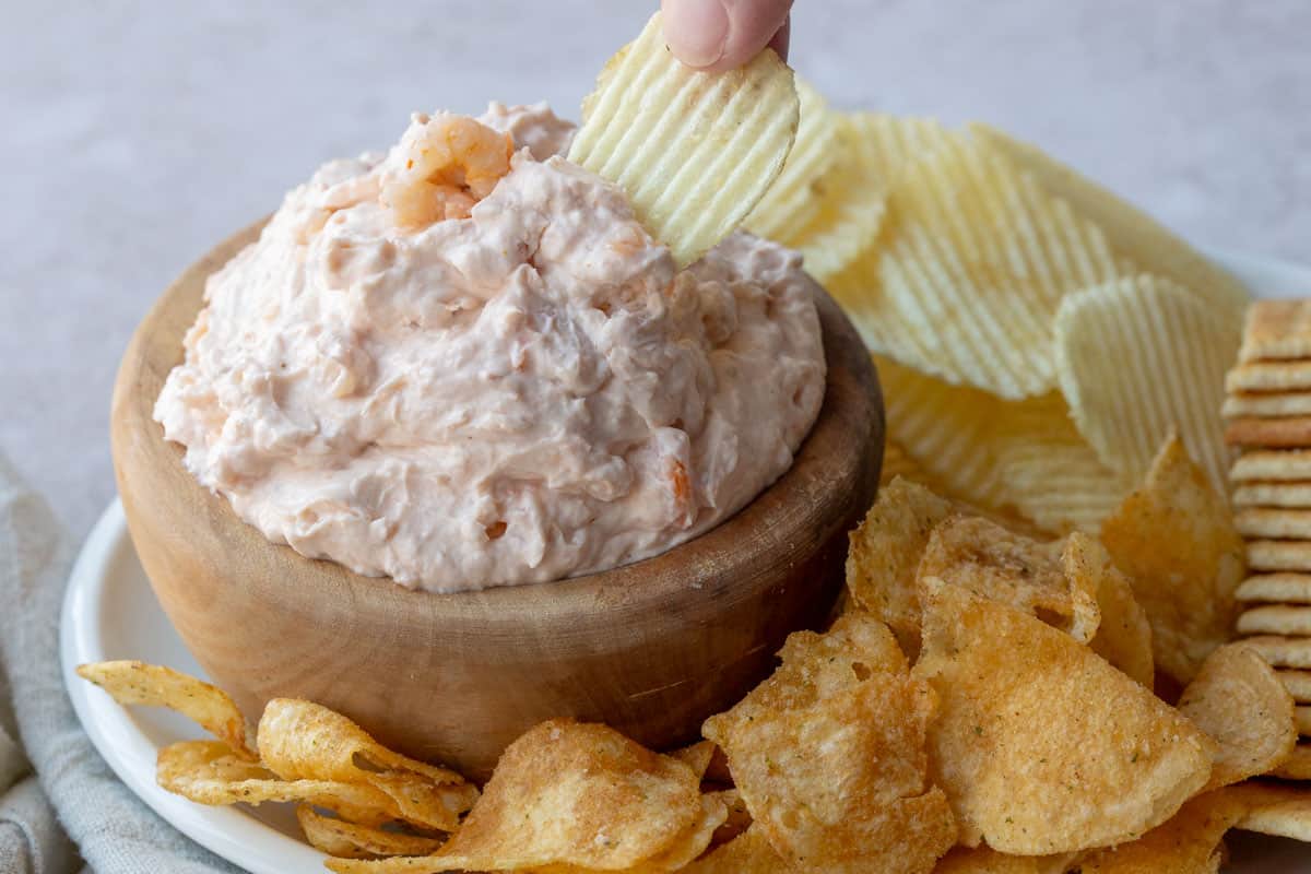 a hand dipping a ruffled potato chip into a bowl of shrimp dip.