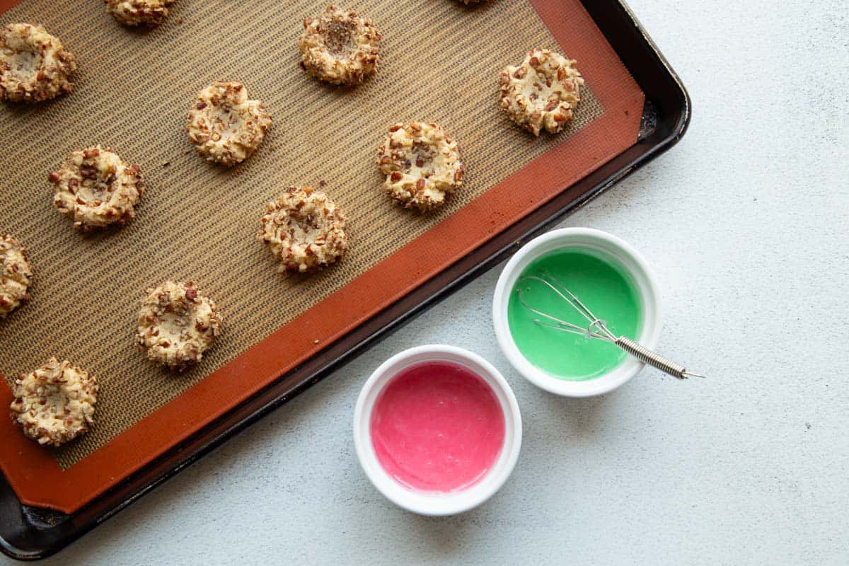 2 small bowls of colored icing next to baked thumbprint cookies.