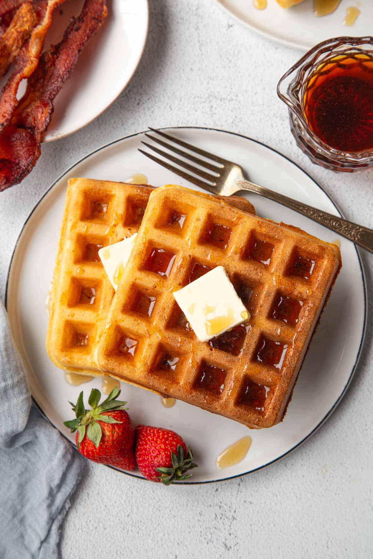 two yeast waffles on a plate with strawberries, a pat of butter, and maple syrup.