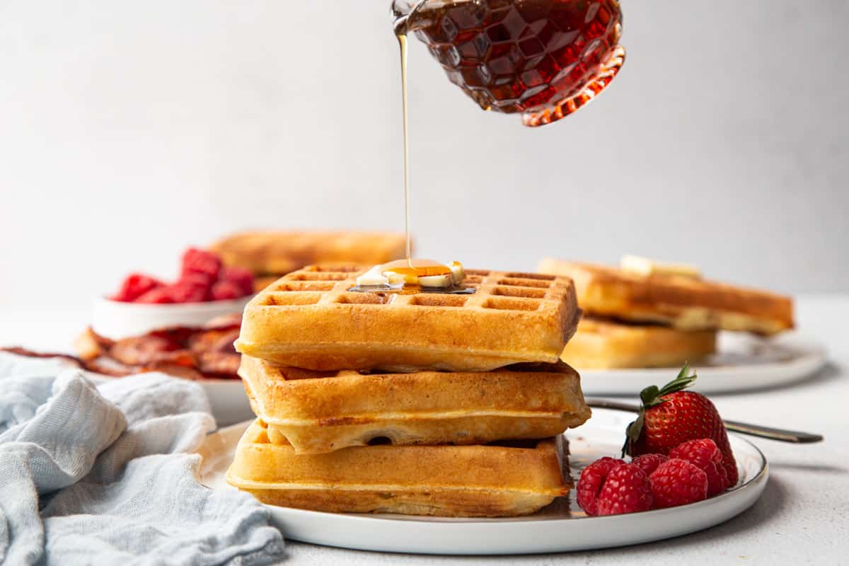 belgian yeast waffles stacked on a plate with a mini pitcher pouring maple syrup on top.