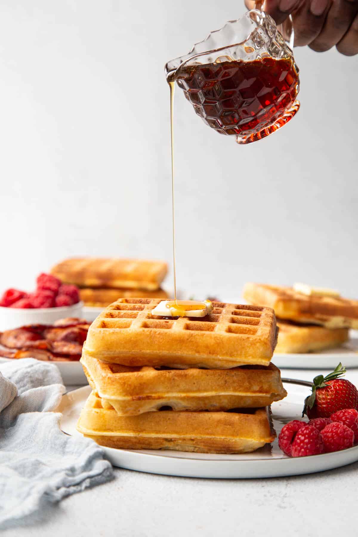 hand pouring maple syrup over a stack of waffles on a plate with raspberries.