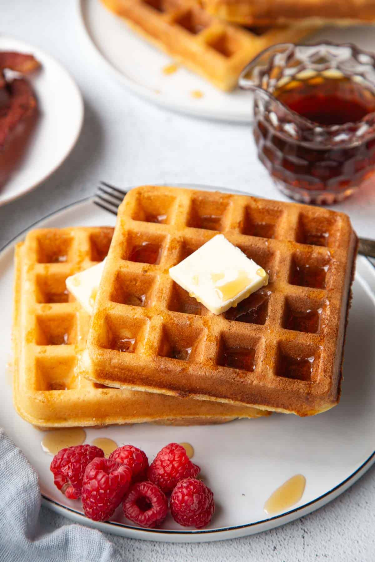 square belgian waffles with butter and syrup on a plate with raspberries.