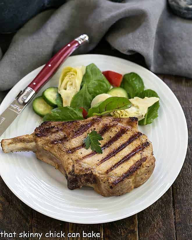 a pork chop on a white plate next to a knife and salad.