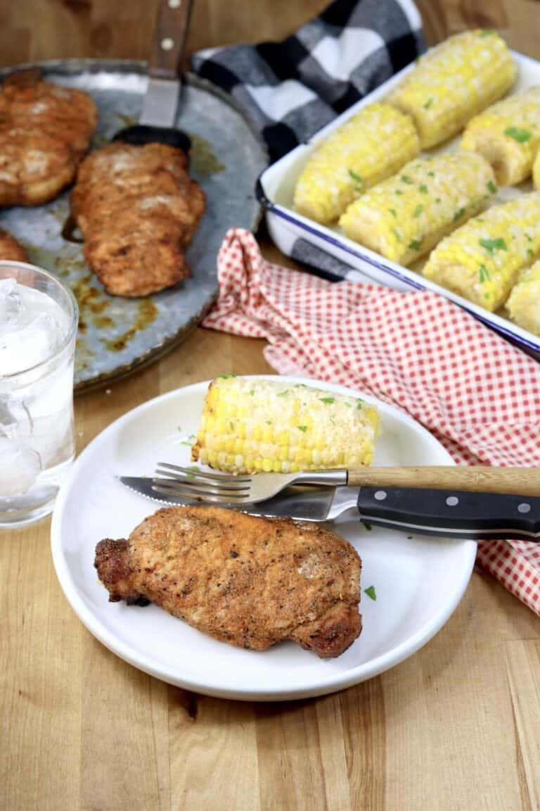a grilled pork chop on a white plate next to grilled corn and silverware.