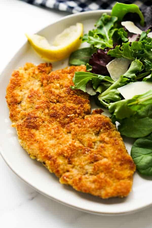 a piece of pork milanese on a white plate next to a green salad.