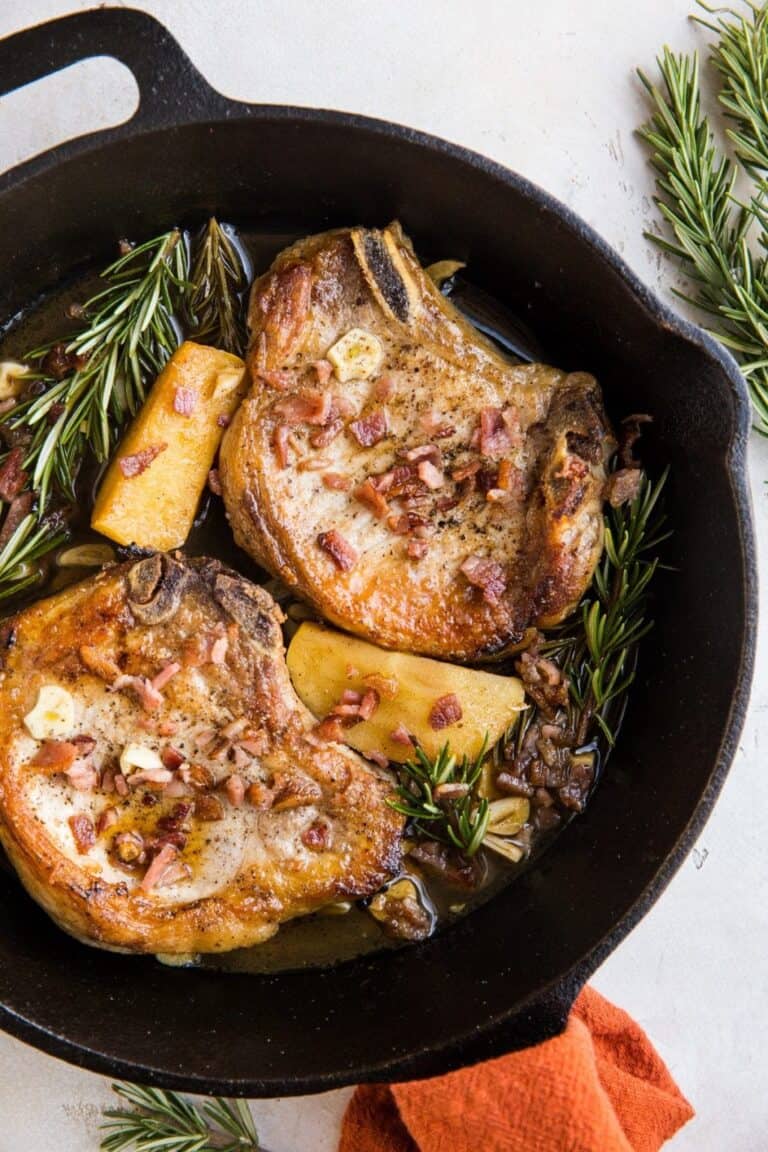 two pork chops in a skillet with rosemary, apples and bacon.