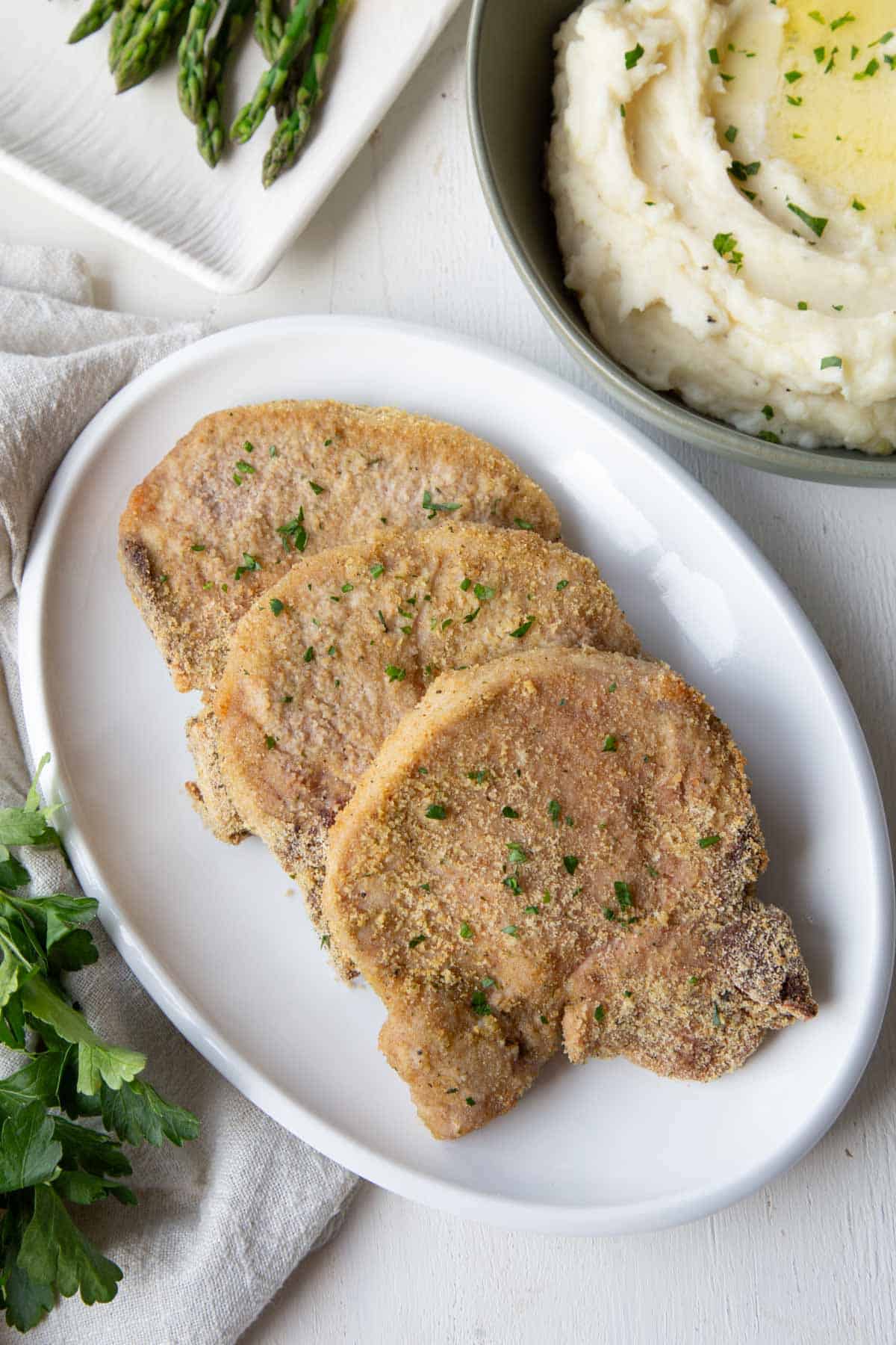 shake and bake pork chops on a platter next to mashed potatoes and asparagus.