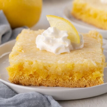 slice of lemon cake with a yellow crust and yellow filling, topped with a dollop of whipped cream and a wedge of lemon.