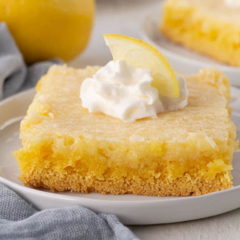 slice of lemon cake with a yellow crust and yellow filling, topped with a dollop of whipped cream and a wedge of lemon.
