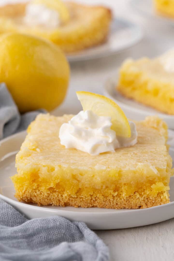 slice of lemon gooey butter cake with a yellow crust, yellow gooey layer, and whipped cream on top.