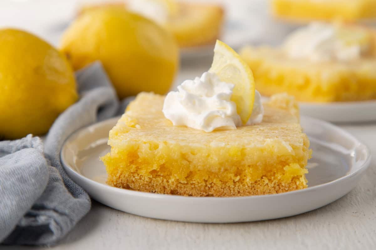 slice of lemon gooey butter cake with a cake mix base and a gooey yellow lemon layer on top, topped with whipped cream and a lemon slice.