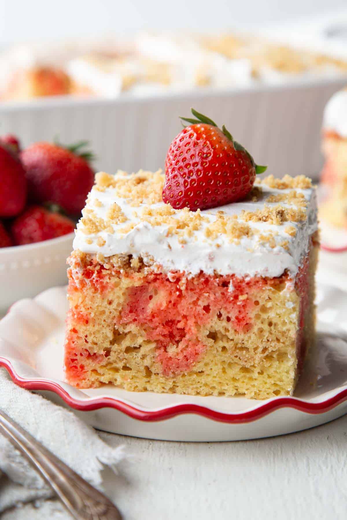 slice of strawberry poke cake with a layer of graham cracker crumbs and nuts, topped with whipped topping and a whole strawberry.