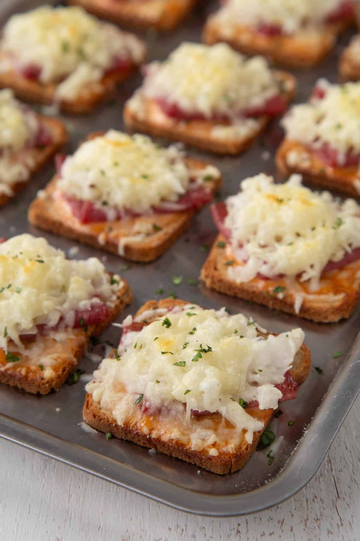 slices of mini rye bread topped with corned beef and swiss cheese on a sheet pan.