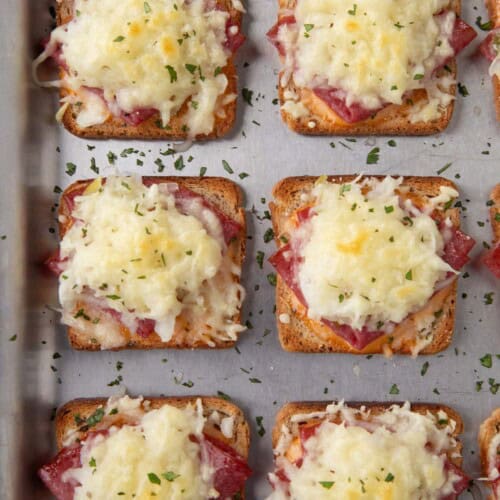 mini reuben sandwiches with a square of toasted rye bread topped with corned beef and melted swiss cheese on a sheet pan.