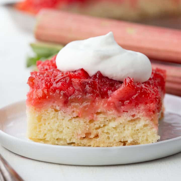 slice of rhubarb upside down cake with white cake on the bottom and a pink rhubarb topping, topped with whipped cream.