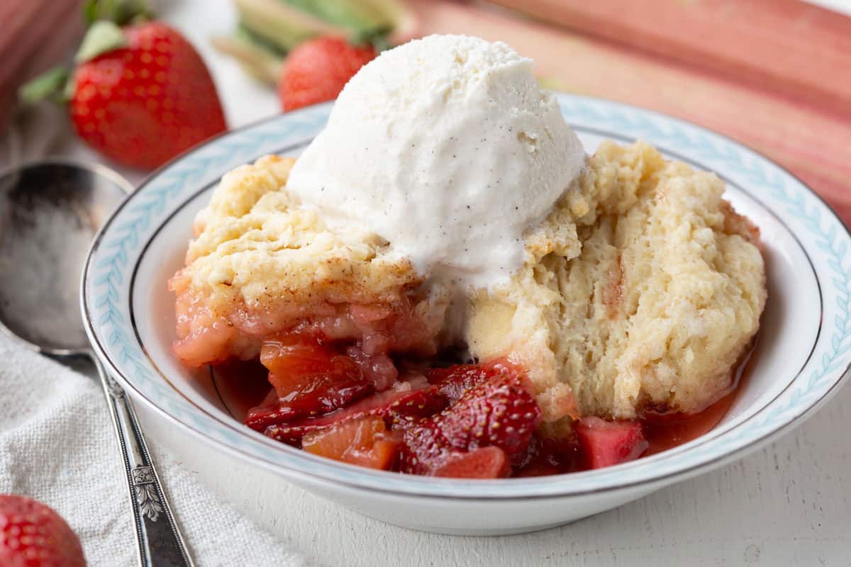 bowl of strawberry rhubarb cobbler with biscuit topping, topped with a scoop of vanilla ice cream.