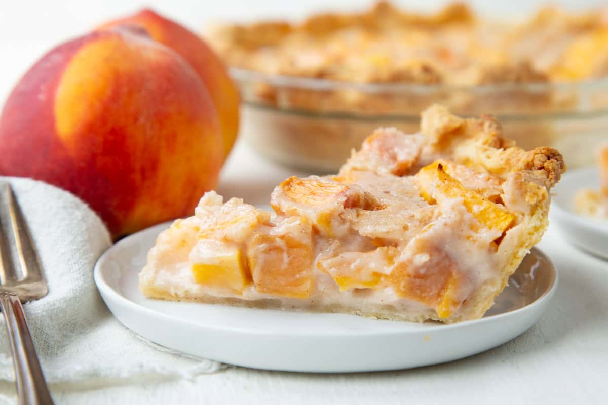 slice of peach cream pie on a white plate next to a fresh peach.