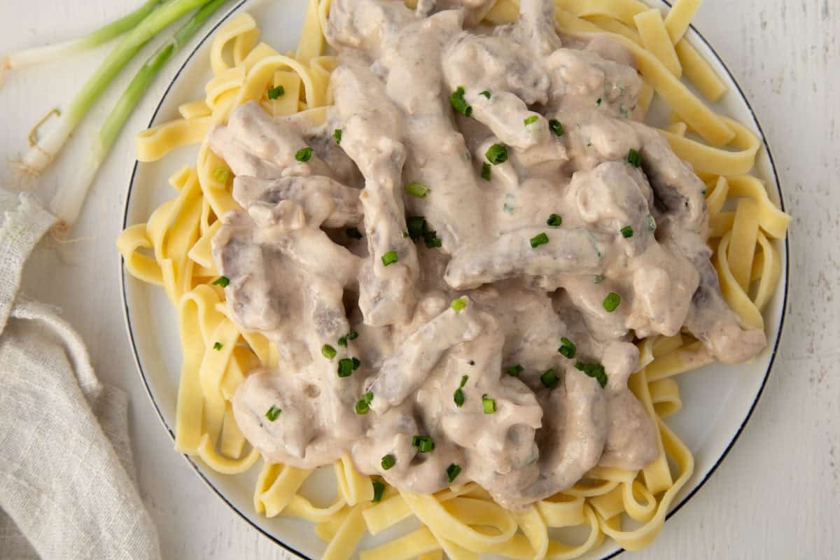 strips of beef in a creamy onion sauce over egg noodles, next to a few green onions.