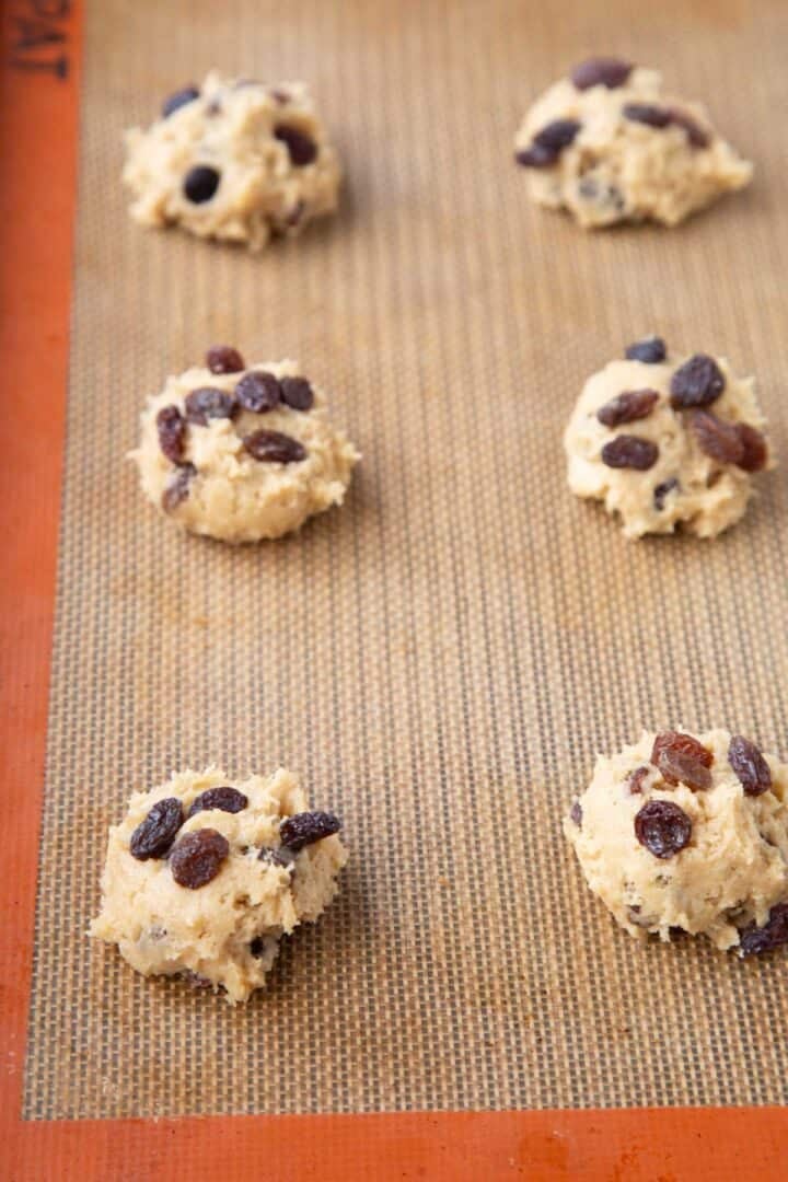 balls of cookie dough topped with raisins on a baking sheet.
